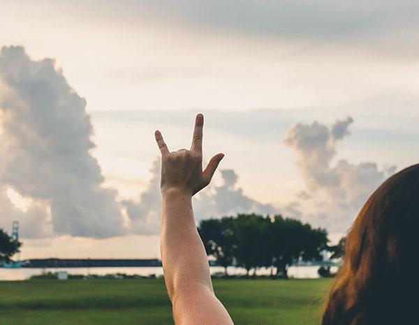 A hand holding up the 'Phins Up' symbol with fingers extended to create a J and a U against a bright sky.